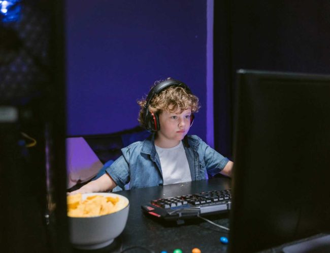 A child wearing headphones intensely playing video games in a dimly lit room with snacks nearby.