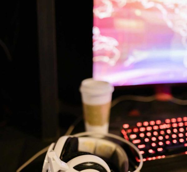 A gaming desk setup with white headphones, keyboard, blurred screen, and coffee cup.